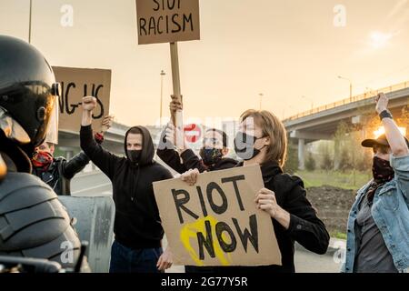 Gruppe von verärgerten Menschen in Masken mit Schildern, die die gleichen Rechte für alle Ethnien fordern, während sie auf der Straße gegen die Bereitschaftspolizei schreien Stockfoto