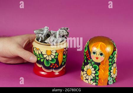 Hand hält russische matryoschka voller Geld. Zerknitterte hundert-Dollar-Scheine fallen aus offenem Holzspielzeug. Symbol für Russland und amerikanisches Geld auf rosa BA Stockfoto