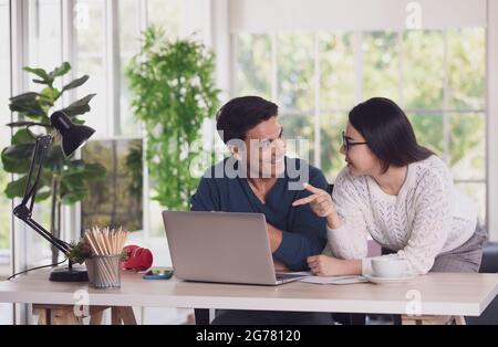 Der kaukasische Mann und die asiatische Frau mischen sich zwischen Rennliebhabern, die über einen Laptop-Notebook-Computer zusammenarbeiten und im Wohnzimmer mit glücklichen und intimen Menschen diskutieren. Neu Stockfoto