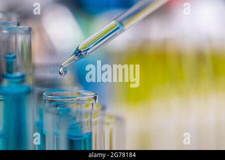 Tropfen blauer Flüssigkeit vom Tropfen zum Reagenzglas im Labor mit heller Farbunschärfe im Hintergrund. Stockfoto