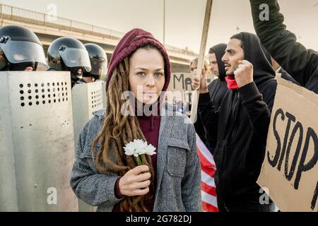Porträt einer verängstigten jungen kaukasischen Frau mit Schrecken, die mit Gänseblümchen zwischen Polizeikräften und Protestierenden bei der Kundgebung stand Stockfoto