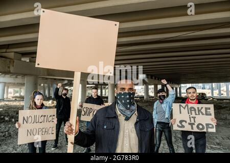 Gruppe von ernsthaften, brutalen jungen Männern mit gemischter Rasse in Bandana auf der Nase, die ein leeres Schild mit der Überschrift der Menge von Aktivisten halten Stockfoto