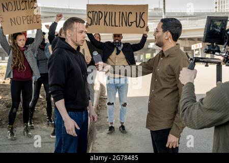 Junger arabischer Journalist in Brillen, der mit gebrochenem Kopf mit einem Aufrührer spricht, während er ihn am Boden interviewt, Menschen mit Schildern im Hintergrund Stockfoto