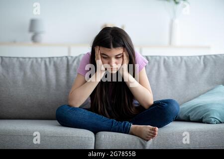 Trauriges indisches Teenager-Mädchen, das zu Hause mit dem Kopf in den Händen auf dem Sofa sitzt und unter mangelnder Kommunikation und Einsamkeit leidet Stockfoto