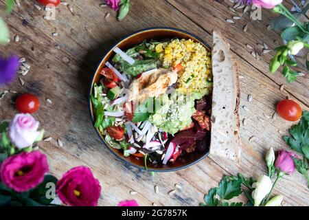 Schüssel mit gesundem Salat mit Avocado, Gemüse und Samen auf einem Holztisch zwischen Blumen Stockfoto