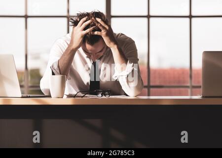 Der junge Geschäftsmann sitzt allein am Schreibtisch und hält seinen Kopf mit den Händen und fühlt sich traurig, ernst und stressig wegen der Arbeitsprobleme im Büro Stockfoto