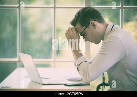 Der junge kaukasische Geschäftsmann sitzt allein im Büro und fühlt sich gestresst und verwendet die Hand, die seine Stirn hält, nachdem er während des wirtschaftlichen Abfeuers abgefeuert wurde Stockfoto