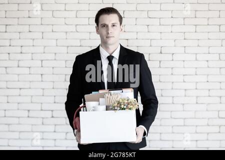 Junger kaukasischer Geschäftsmann in schwarzem Anzug mit einem traurigen, gestressten und unglücklichen Gesicht, das einen Papierkarton mit Habseligkeiten hält, die im Get Fi im Büro gesammelt wurden Stockfoto