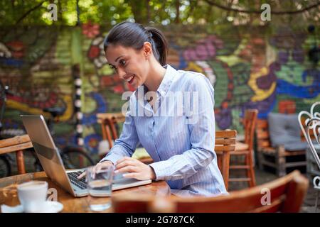 Porträt einer jungen, schönen kaukasischen erwachsenen Frau, die im Café im Freien sitzt, lächelt, auf ihren Laptop schaut, schreibt. Online-Kommunikationskonzept Stockfoto