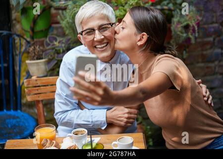 Ein junges Mädchen macht in freundlicher Atmosphäre in der Bar ein Selfie, während sie ihrer älteren Freundin einen freundlichen Kuss gibt. Freizeit, Bar, freundlich Stockfoto