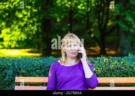 Unschärfe kaukasisch blonde Frau reden, sprechen am Telefon draußen, im Freien. 40-jährige Frau in lila Bluse im Park auf der Bank. Erwachsene weibliche Erbsenw Stockfoto