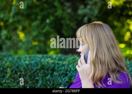 Unschärfe kaukasisch blonde Frau sprechen auf dem Telefon draußen, draußen. 40er Jahre alte Frau in lila Bluse im Park, Profil. Seriöse Person, die mobil verwendet Stockfoto