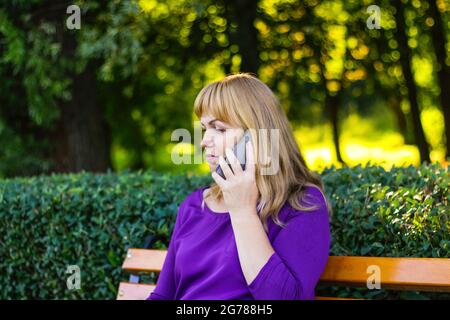 Unschärfe kaukasisch blonde Frau reden, sprechen am Telefon draußen, im Freien. 40-jährige Frau in lila Bluse im Park auf der Bank. Erwachsene Person usi Stockfoto