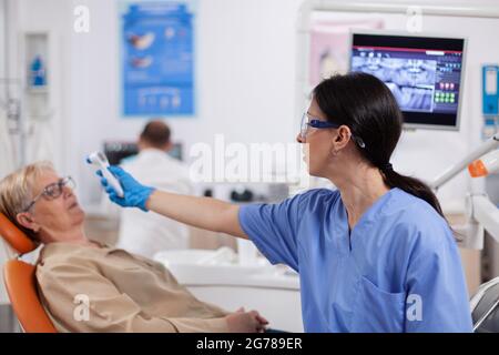 Zahnarzthelferin Messung Senior Frau Körpertemperatur mit Thermometer während der Konsultation. Medizinische Facharzt in der Zahnklinik unter Patienten Temperatur mit digitalen Gerät. Stockfoto