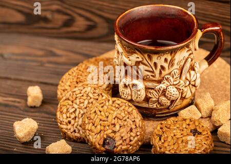 Hausgemachte Kekse mit Sonnenblumenkernen und Rosinen, Stücke von braunem Rohrzucker und ein Tonbecher Tee aus Steingut auf einem Holztisch. Nahaufnahme des selektiven Fokus Stockfoto