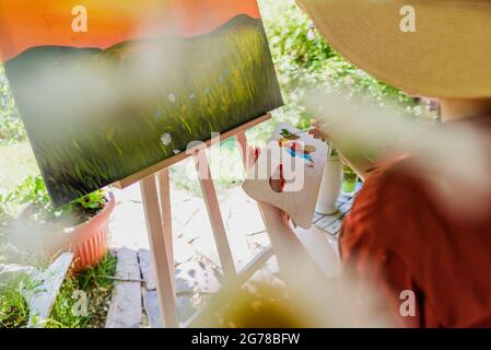 Junge Künstlerin arbeitet an ihrer Kunst Leinwand Malerei im Freien in ihrem Garten. Achtsamkeit, Kunsttherapie, Kreativität und kreative Hobbys Konzept. Stockfoto