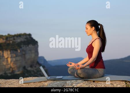 Seitenansicht eines asiatischen Yogis, der Yoga mit einem Laptop in den Bergen bei Sonnenuntergang lernt Stockfoto