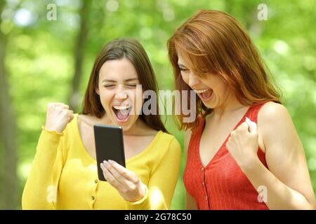 Zwei aufgeregte Frauen überprüfen den Inhalt von Smartphones in einem grünen Park oder Wald Stockfoto