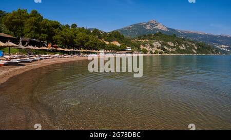 Ionische Inseln, Zakynthos, südlich der Stadt Zakynthos, Banana Beach, halbrunde Bucht, Sandstrand, aufgereihte Sonnenliegen mit Sonnenschirmen, Meer grün-blau, himmelblau, Berg im Hintergrund Stockfoto