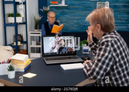 Fröhliche ältere Frau winkt während eines Online-Anrufs mit Verwandten vor der Webcam. Glückliche Großmutter, die im Rahmen von Online-Video grüßt, konfenzere mit Familie aus dem Wohnzimmer. Stockfoto