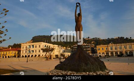 Zakynthos, Zakynthos-Stadt, Solomos-Platz, Morgenlicht, Bronzestatue der Freiheit, Weitwinkelansicht, Statue und Sockel rechts im Bild, Solomos-Platz mit Gebäuden im Hintergrund, weiße Wolken am blauen Himmel Stockfoto