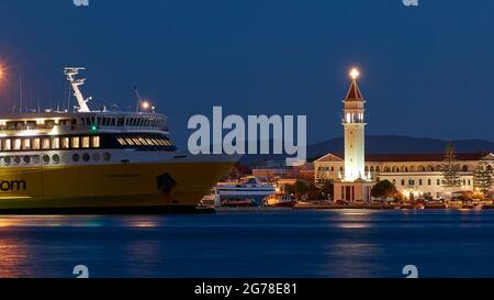 Zakynthos, Zakynthos Stadt, Nachtaufnahme, Abendaufnahme, Hafen, Vorderteil der Zante-Fähre in der mittleren Entfernung, im Vordergrund blaues Wasser, beleuchtete Agios Dionysios-Kirche direkt hinter der Fähre, blauer Nachthimmel, festlich beleuchteter Kirchturm Stockfoto