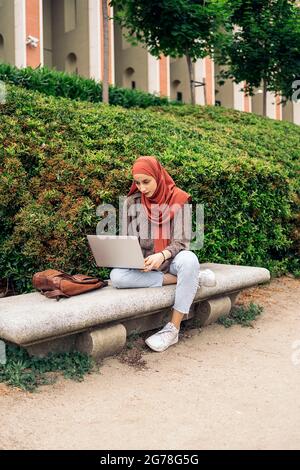 Junge muslimische Frau mit Hijab sitzt im Park und benutzt ihren Computer. Stockfoto