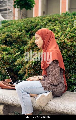 Schöne junge muslimische Frau mit Hijab, die im Park sitzt und ihren Computer benutzt. Stockfoto
