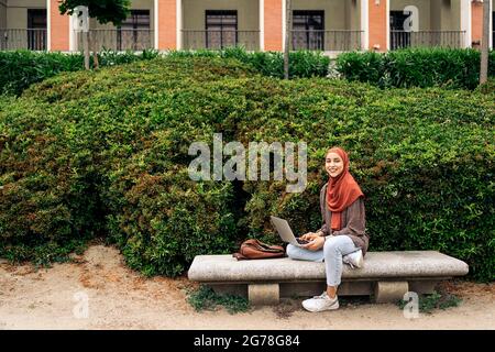 Glückliche muslimische Frau, die im Park sitzt und ihren Laptop benutzt. Sie lächelt und schaut in die Kamera. Stockfoto