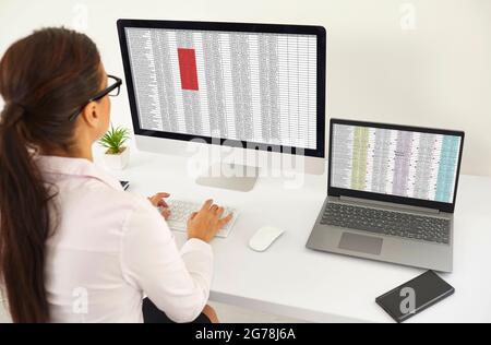 Frau, die am Schreibtisch sitzt und mit Datenlisten auf Desktop- und Laptop-Computern arbeitet Stockfoto