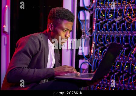 Konzentrierter junger afroamerikanischer IT-Ingenieur, der mit blauen Kabeln in der Nähe des Serverschranks hockt und Touchpad auf dem Laptop verwendet Stockfoto