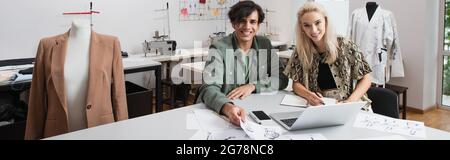 Junge Designer lächeln der Kamera in der Nähe von Gadgets und Skizzen am Arbeitsplatz, Banner Stockfoto
