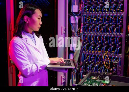 Seriös beschäftigte junge asiatische Netzwerkingenieurin im Labormantel, die mit einer an den Server angeschlossenen Leiterplatte auf dem Wagen steht und Daten auf einem Laptop analysiert Stockfoto