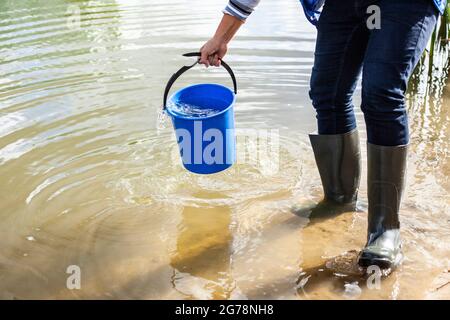 Eimer mit Wasser. Ein Mann zieht Wasser aus einem Teich. Wasser aus einem Reservoir nehmen. Eimer in der Hand. Der Typ hält das Gewicht. Stockfoto