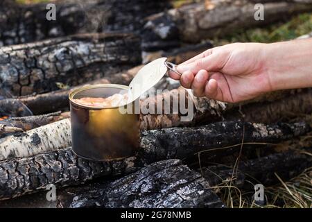 Zinndose auf dem Feuer. Aufwärmen von Fleisch bei einem Picknick. Fleischkonserven aus Holzkohle. Der Tourist bereitet das Mittagessen zu. Stockfoto