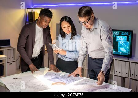 Team von konzentrierten interrassischen Automobilingenieuren, die im modernen Büro mit Neonbeleuchtung an einem Tisch stehen und Autoskizzen besprechen Stockfoto