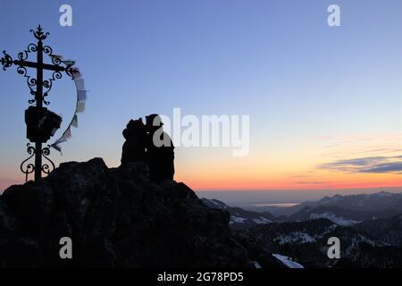 Junges Paar tief umarmen, küssen, genießen den Sonnenaufgang und den Blick vom Auerspitz (1811m) in Richtung Chiemgau, rechts vom Paar das Wildalpjoch (1725m) hinter dem Chiemsee, die Kampenwand (1668m) ganz rechts, stimmungsvoll, Oberbayern, Bayern, Deutschland , Bayerische Alpen, Mangfallgebirge Stockfoto