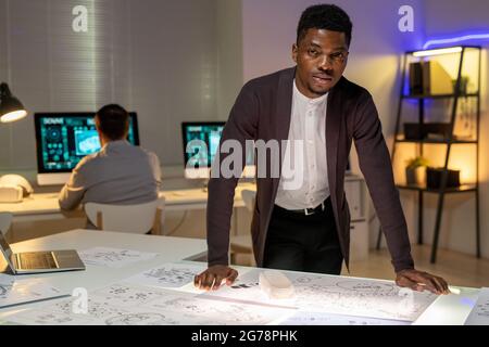 Porträt eines ernsthaften afroamerikanischen Designers, der sich mit Skizzen im dunklen Büro auf den Tisch lehnte und in einem Automobilhersteller arbeitete Stockfoto