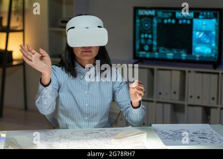 Junge asiatische Frau, die mit der Hand durch Designbilder blättert, während sie mit einer VR-Brille an der Außenseite des Autos arbeitet Stockfoto