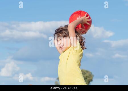 Kleiner Junge mit langen Haaren und gelbem T-Shirt spielt an einem sonnigen Sommertag mit seiner roten Kugel auf einem Feld Stockfoto