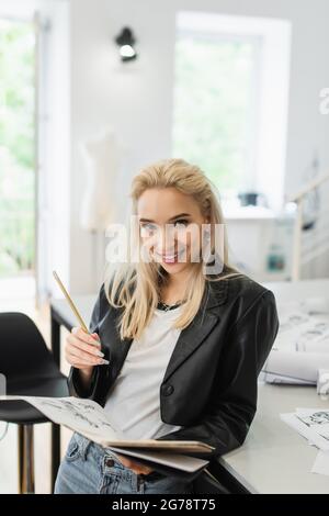 Fröhliche Modedesignerin mit Notizbuch, Bleistift und Zeichnung, die in einem atelier die Kamera anschaut Stockfoto