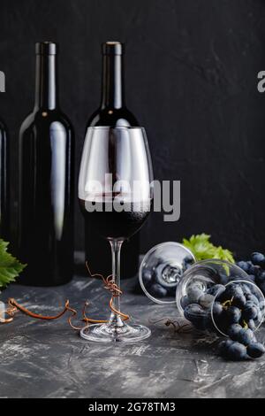 Weinkomposition auf schwarzem Steintisch. Rotweinflaschen, Weintrauben mit Blättern und Reben auf dunklem rustikalem Betonhintergrund. Weinglas mit Rot Stockfoto