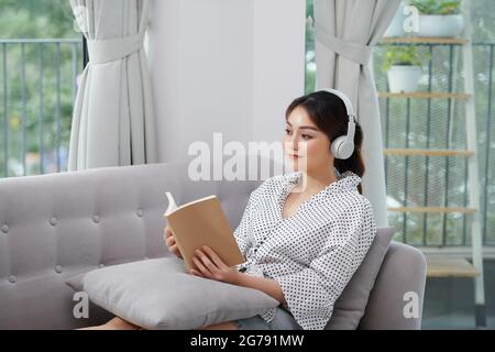 Eine Frau liest ein Buch und hört Musik, während sie auf einem grauen Sofa sitzt. Stockfoto