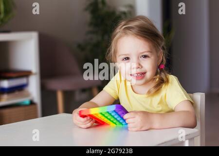 Kleine blonde Mädchen Kinder spielen mit neuen Trend sensorische Spielzeug Regenbogen Pop it. Das Mädchen lächelt und schaut auf die Kamera Stockfoto