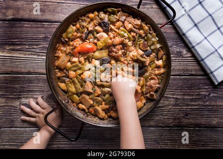 Kinderhand versucht eine Paella. Holztisch mit einer traditionellen hausgemachten Paella.Vegane Paella Stockfoto