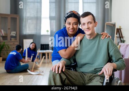 Glückliche junge Mischrasse weibliche Freiwillige umarmt behinderten Mann im Rollstuhl Stockfoto