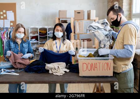 Drei Freiwillige in Masken und Handschuhen sortieren Spendenkleidung Stockfoto