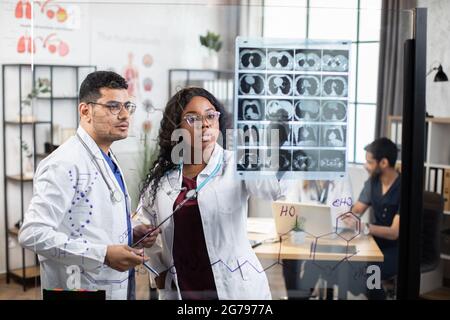 Zwei international kompetente Ärzte diskutieren die Ergebnisse der Röntgen-mrt-Untersuchung. Afrikanische Frau und muslimischer Mann stehen in der Nähe einer Glaswand und schauen sich den Tomographie-Scan an Stockfoto