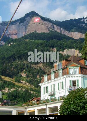 Vierwaldstättersee, Schweiz: August 10 2005: Ein altes weißes Hotel mit grünen Fensterläden vor einem großen Berg mit einer Felswand, auf der eine Schweizer Flagge ca. Stockfoto