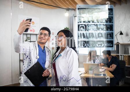 Lächelnde afrikanische Frau und indischer Mann im weißen Laborkittel, die Selfie auf dem Smartphone machen. Zwei medizinische Mitarbeiter, die nach einem internationalen Treffen Fotos machen. Blick durch die Glaswand Stockfoto